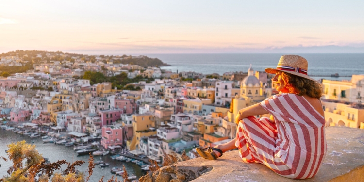 Side view of positive mature female traveler in striped dress and hat enjoying amazing seascape during sunset in Procida island.